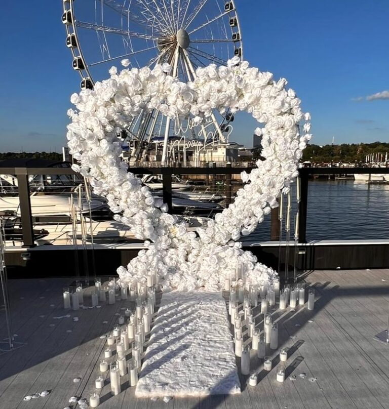 A white rose heart arch with candles and a Will You Marry Me sign set up on a marina deck with a Ferris wheel in the background.