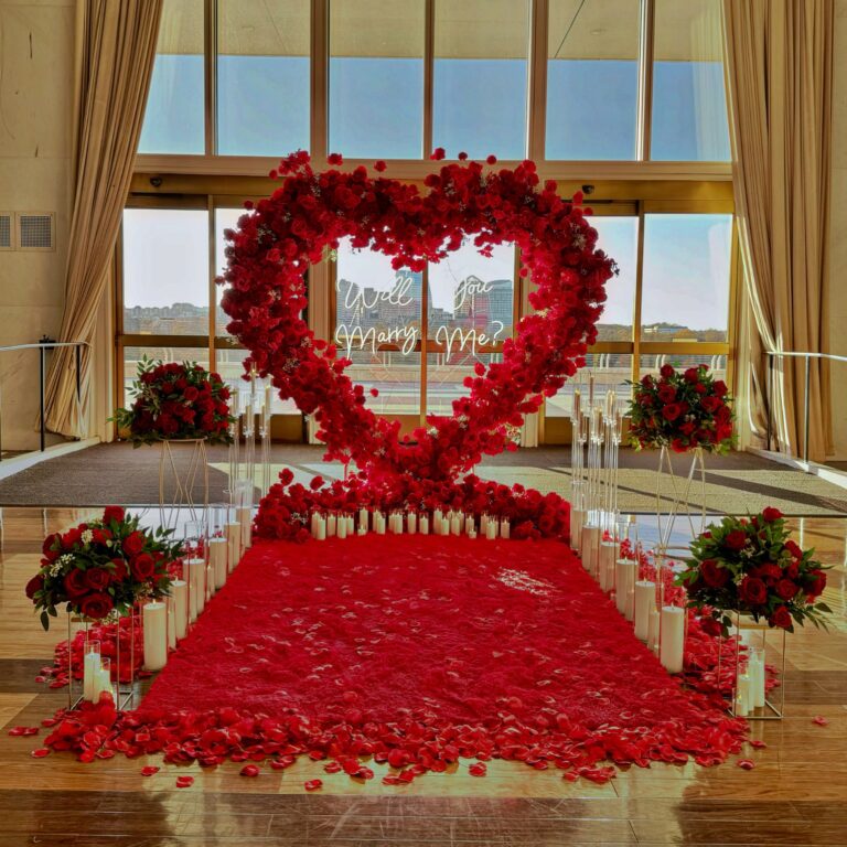 Luxury indoor marriage proposal setup with a large heart shaped red rose arch, glowing “Will You Marry Me” sign, candlelit aisle, and rose petal carpet inside an elegant ballroom overlooking the city.